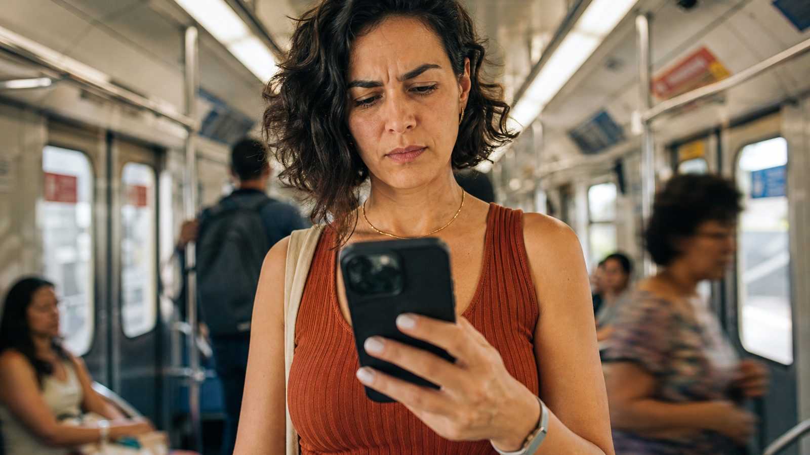 Mulher de uns 35 anos no metrô, atenta ao celular, com a sobrancelha erguida de quem confere uma informação, desconfiada mas calma