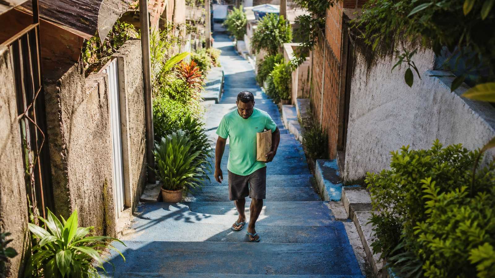 Homem de camiseta verde subindo com calma uma escadaria de rua com degraus pintados de azul, num bairro ensolarado de manhã