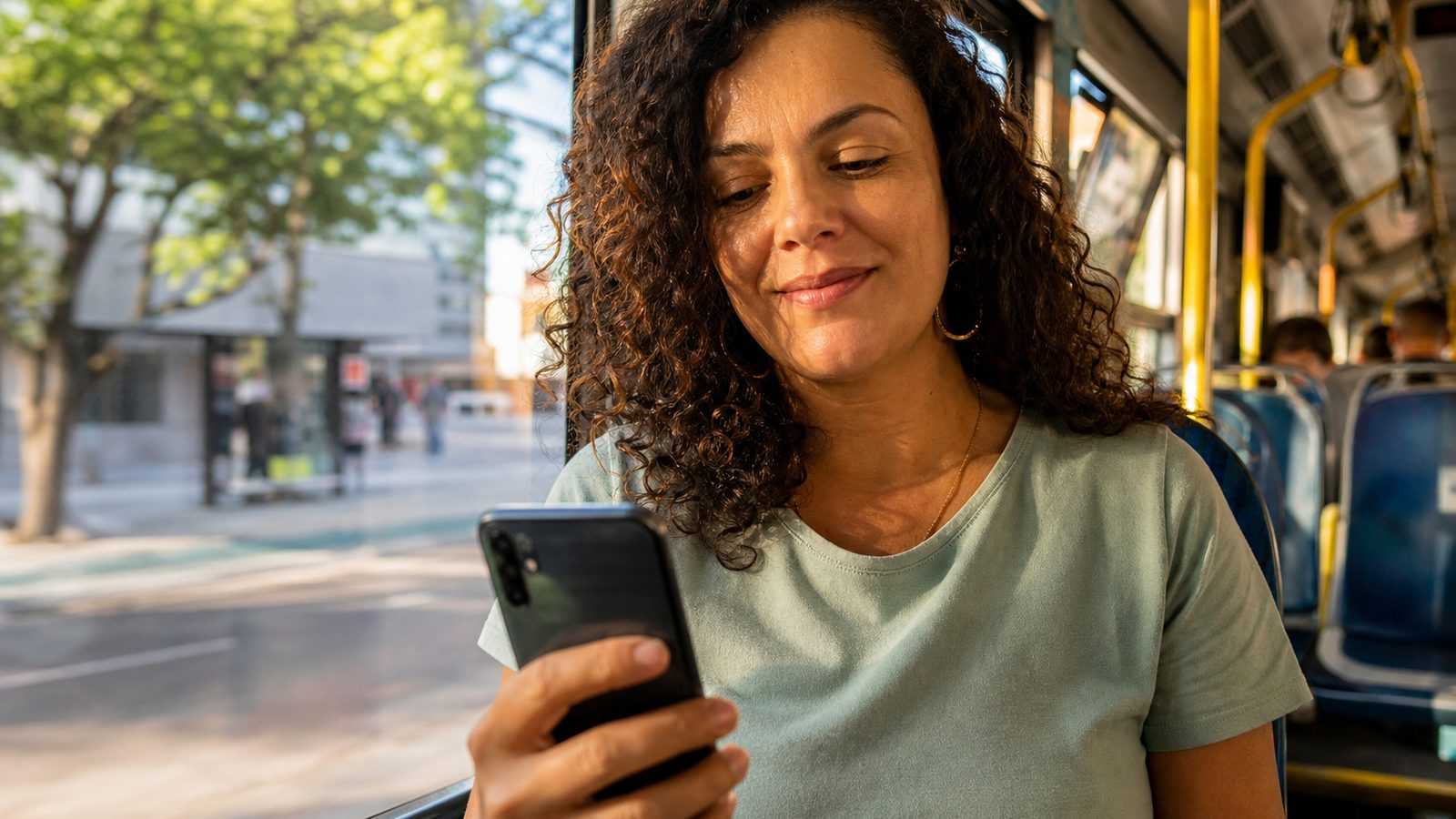 Mulher olhando o celular com expressão tranquila, sentada perto da janela de um ônibus no fim da tarde
