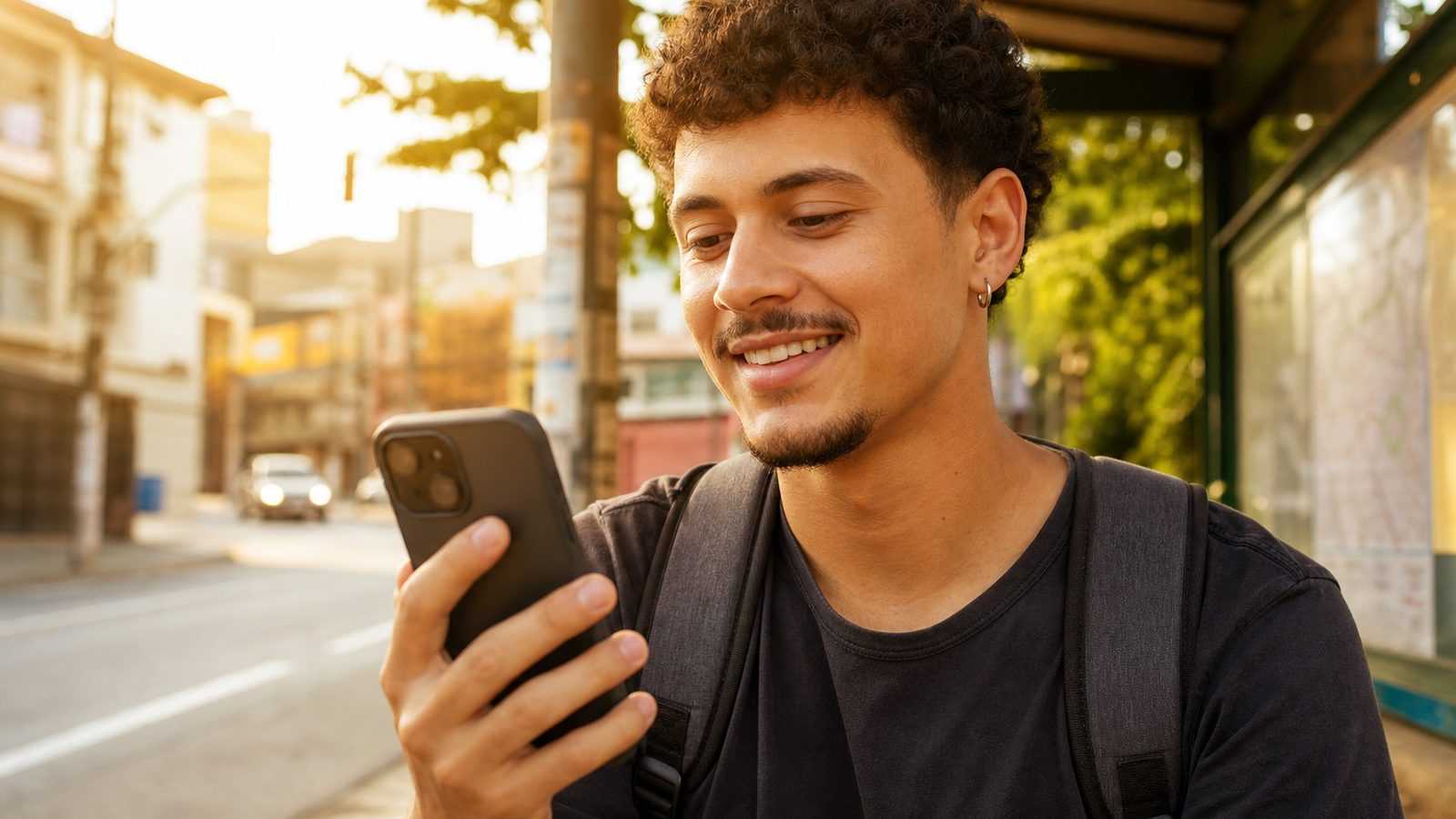 Jovem de uns 25 anos no ponto de ônibus, olhando o celular com curiosidade e alívio, sob a luz do fim de tarde